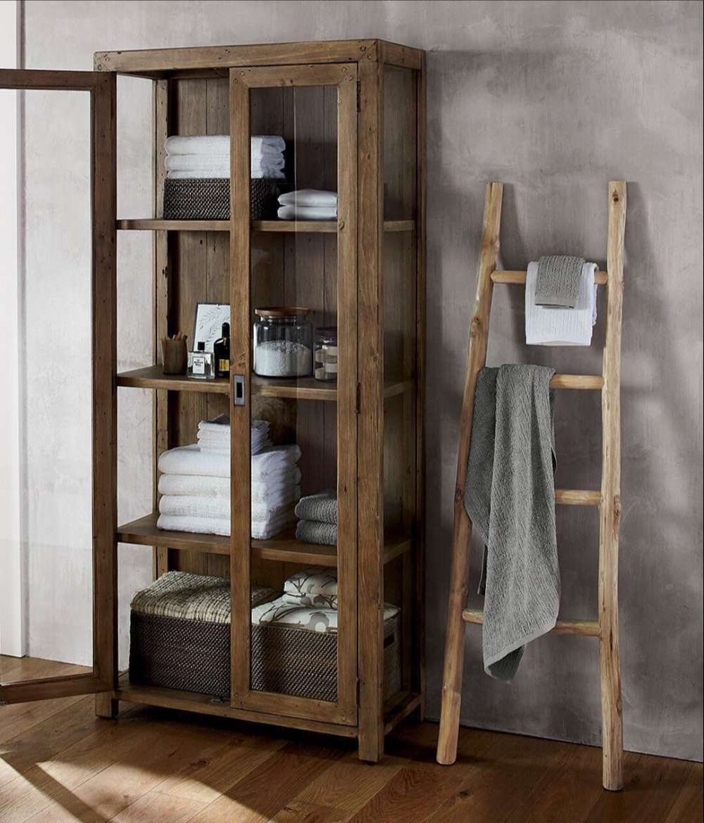 Bathroom storage setup featuring Crate & Barrel Morris 36" Chocolate Brown Wood and Glass Bookcase Cabinet with towels and baskets