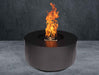 Black Warming Trends AON Steel Circular Fire Pit Table is shown burning strongly in a staged setting with a gray floor and dark wall background