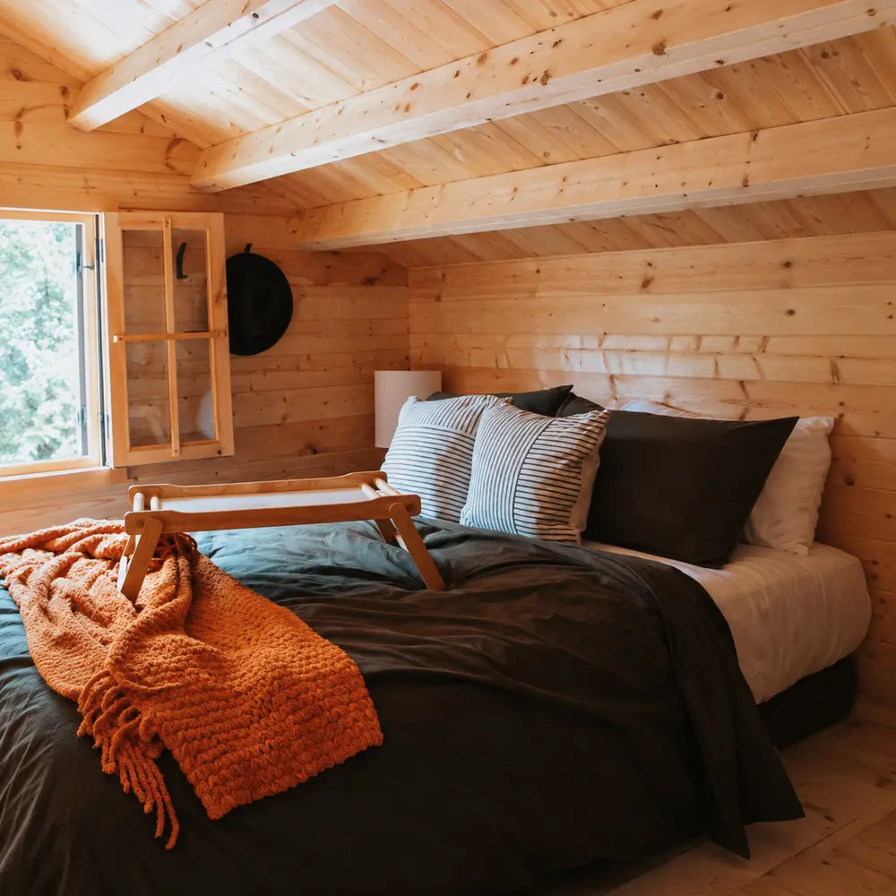 Warm upper-level sleeping nook in the BlueWater Loft 160 38mm with dark bedding, striped pillows, an orange blanket, and a sloped pine ceiling.