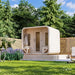 Side-angle view of the Sawmill Structures Box Sauna surrounded by green trees, purple flowers, lawn, and a horizontal wooden privacy fence.