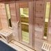 Glass-fronted entrance to the Sawmill Structures Box Sauna with a wooden handle, narrow side windows, porch benches, and interior seating visible through the door.