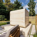 Rear view of the Sawmill Structures Box Sauna showing its smooth windowless light-wood wall, rounded corners, raised platform, and garden surroundings.