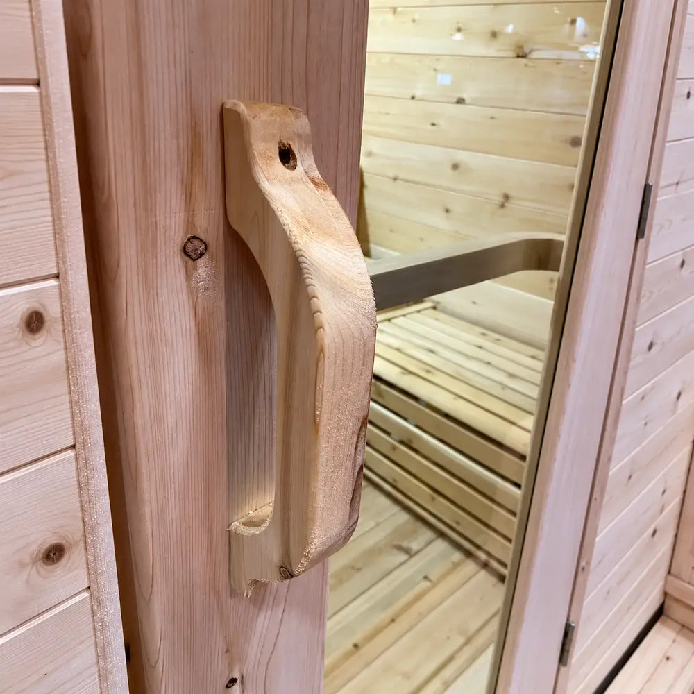 Close-up of the sculpted wooden door handle on the Sawmill Structures Box Sauna with the glass panel and slatted interior bench visible behind it.