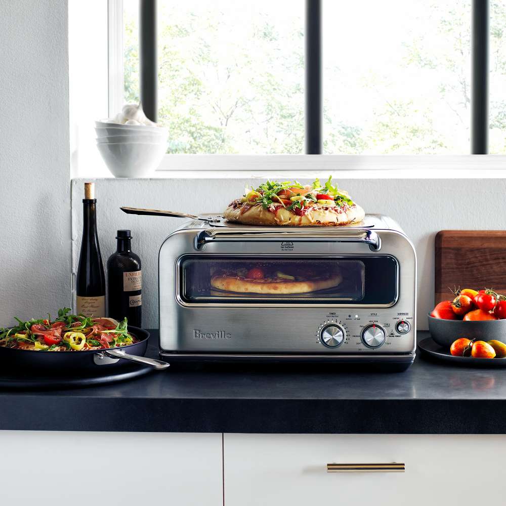 Breville® Smart Oven® Pizzaiolo Pizza Oven sits on a bright kitchen counter with pizza inside and fresh ingredients nearby