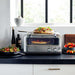 Breville® Smart Oven® Pizzaiolo Pizza Oven sits on a bright kitchen counter with pizza inside and fresh ingredients nearby