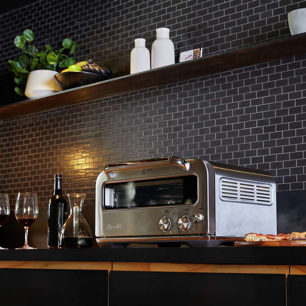 Breville® Smart Oven® Pizzaiolo Pizza Oven sits on a dark kitchen counter beside pizza, wine glasses, and bottles