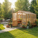 Bright garden scene with sunlight highlights the 8x12 Outdoor Living Today Western Red Cedar Greenhouse Kit next to outdoor seating