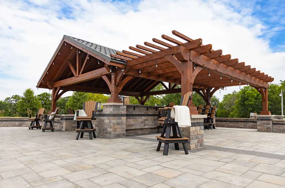 Brookside Timber Frame Combolan Estate Pavilion features a spacious patio with elegant stone flooring, high-top seating, and an inviting outdoor ambiance