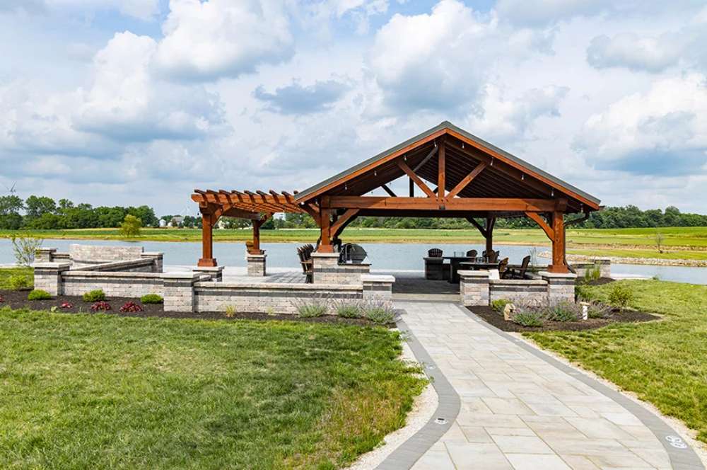 Brookside Timber Frame Combolan Estate Pavilion features a stunning open-air wooden structure with a gabled roof, set beside a peaceful lake with lush greenery