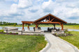 Brookside Timber Frame Combolan Estate Pavilion features a stunning open-air wooden structure with a gabled roof, set beside a peaceful lake with lush greenery