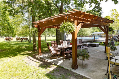 Brookside Timber Frame Fern Shade Pergola covers an outdoor dining area on a concrete patio, surrounded by trees and open grassy land with horses in the distance