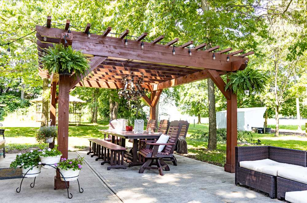 Brookside Timber Frame Fern Shade Pergola frames a spacious patio dining area with surrounding greenery, potted flowers, and comfortable seating