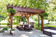 Brookside Timber Frame Fern Shade Pergola frames a spacious patio dining area with surrounding greenery, potted flowers, and comfortable seating