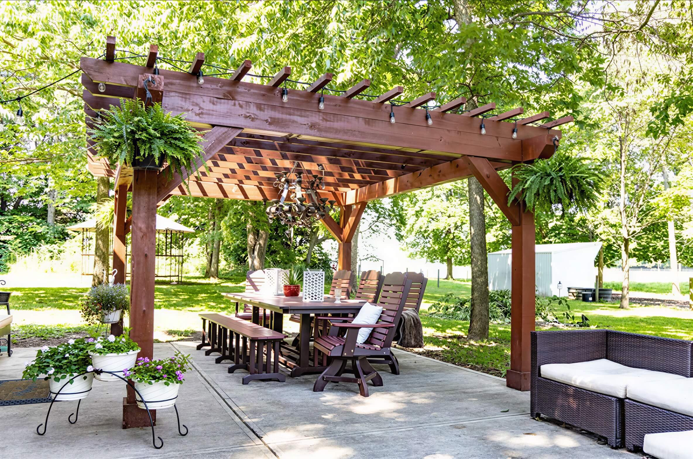 Brookside Timber Frame Fern Shade Pergola frames a spacious patio dining area with surrounding greenery, potted flowers, and comfortable seating