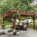 Brookside Timber Frame Fern Shade Pergola stands prominently over a patio table, with hanging plants and soft lighting set against a leafy backyard
