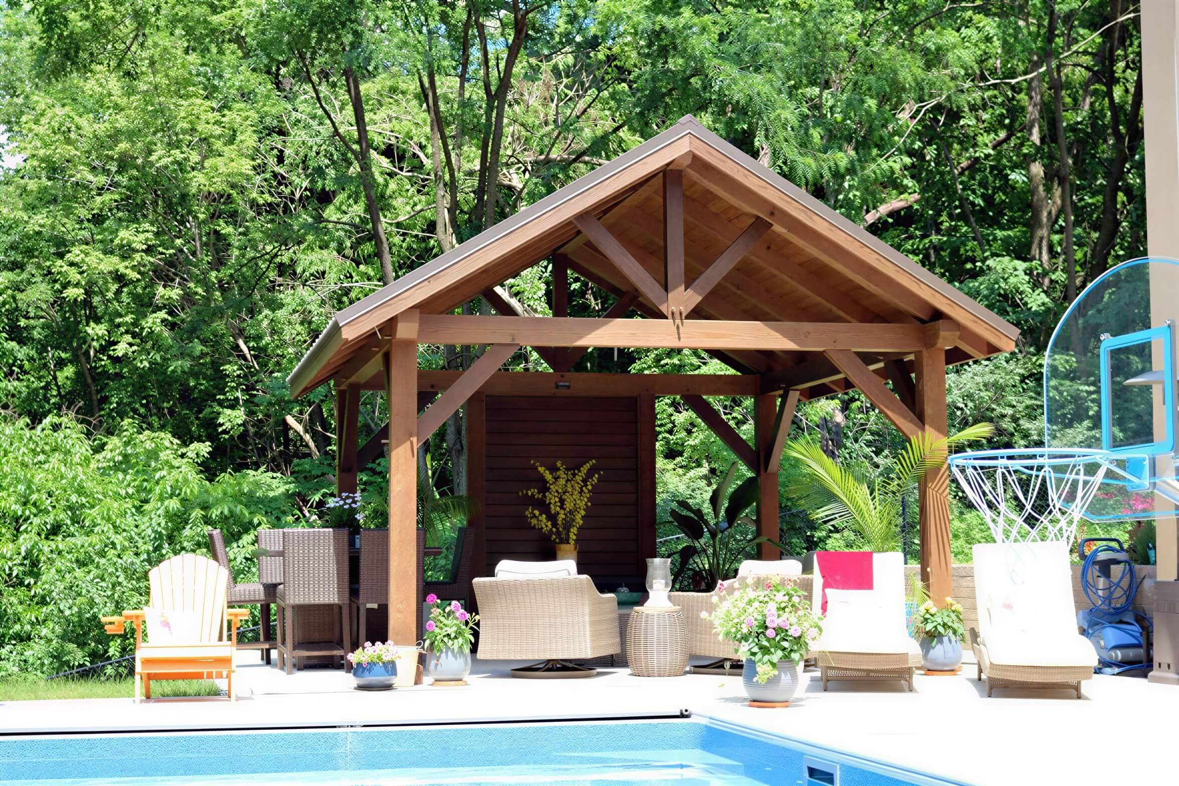 Brookside Timber Frame Forest Retreat Pavilion shelters a relaxed patio setup with wicker chairs, lounge seating, and poolside decor in a bright, sunny backyard
