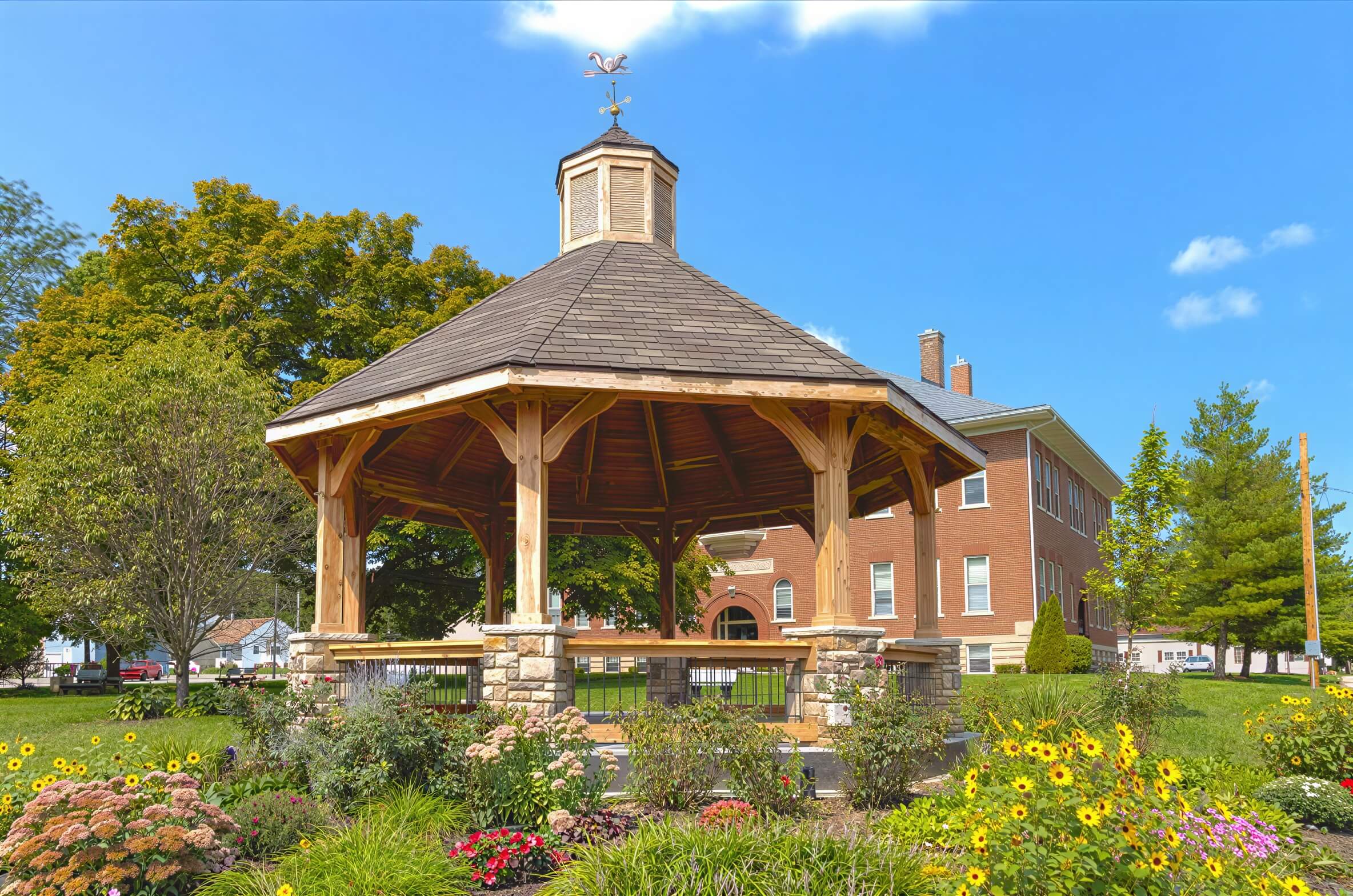 Brookside Timber Frame Lakeside Majestic Pavilion on a sunny day with a detailed roof and blooming garden around it
