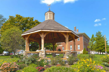 Brookside Timber Frame Lakeside Majestic Pavilion on a sunny day with a detailed roof and blooming garden around it