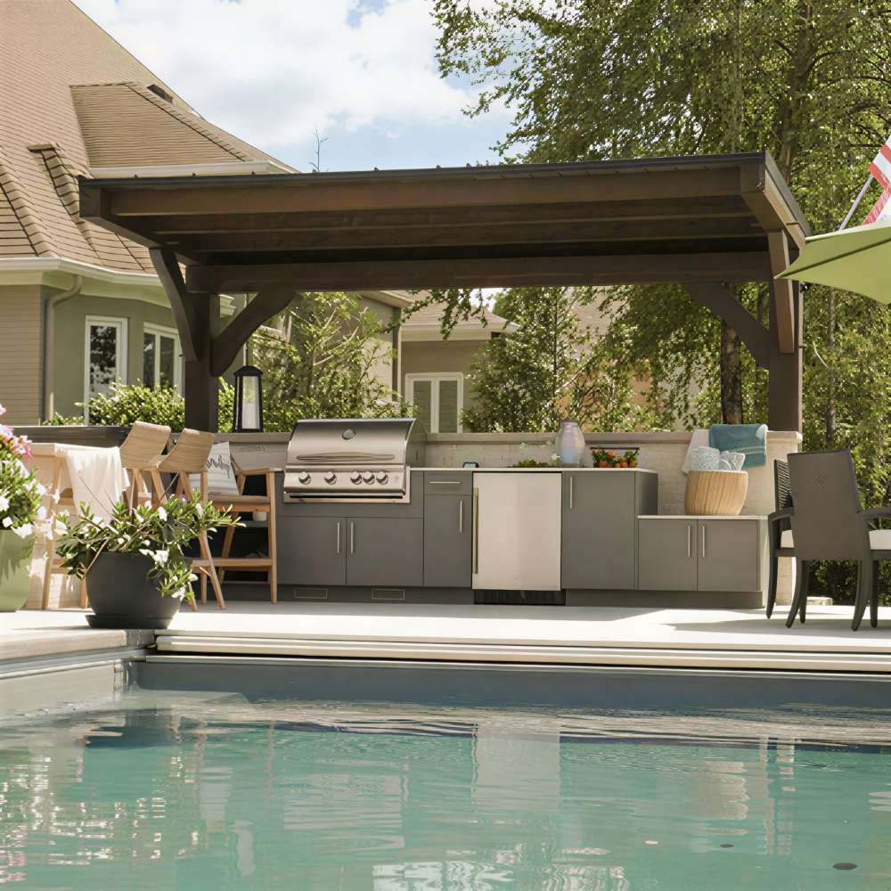 Brookside Timber Frame Ledgefield Pavilion shades a backyard outdoor kitchen with a grill, bar seating, and cabinets beside a clear swimming pool