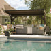 Brookside Timber Frame Ledgefield Pavilion shades a backyard outdoor kitchen with a grill, bar seating, and cabinets beside a clear swimming pool