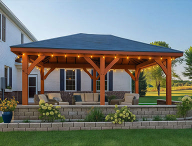 Brookside Timber Frame Timber Crest Pavilion stands in a backyard with a wooden structure, cozy seating underneath, and warm string lights glowing along the roof at dusk