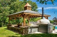 Brookside Timber Frame Forest Melody Pavilion stands tall in a backyard oasis, featuring an elegant wooden structure, a cupola on top, and an outdoor kitchen setup