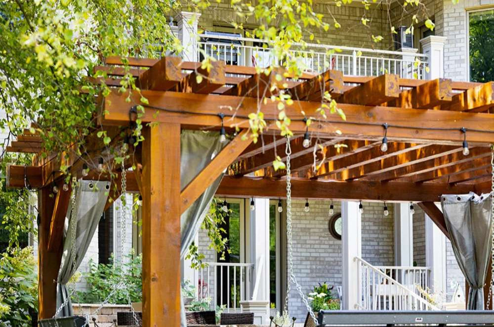 Brookside Timber Frame Highlan Oasis Pergola features a stunning wooden structure with thick beams, decorative braces, and string lights