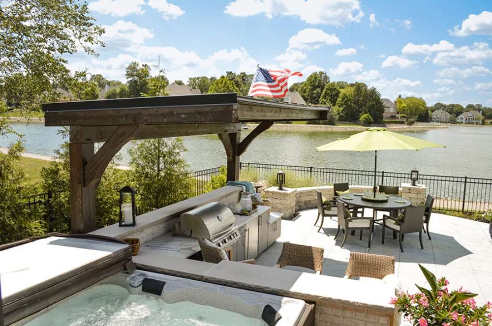 Brookside Timber Frame Ledgefield Pavilion adds rustic elegance to an outdoor living space, complete with a grill, seating area, and serene lake views