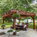 Brookside Timber Frame Shadelan Retreat Pergola creates a charming and functional outdoor oasis, featuring classic wooden furniture, soft lighting, and natural greenery