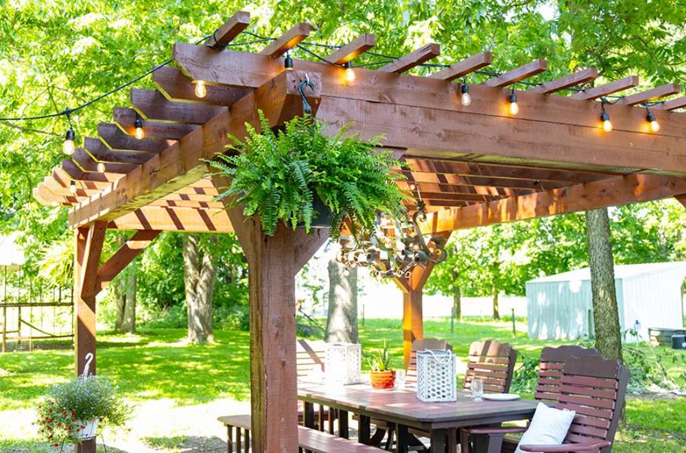 Brookside Timber Frame Shadelan Retreat Pergola stands elegantly over a wooden dining set, adorned with hanging ferns, string lights, and a rustic chandelier