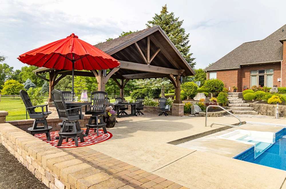 Brookside Timber Frame Timber Retreat Pavilion is a stunning wooden structure by a pool with elegant black patio furniture and a bold red umbrella