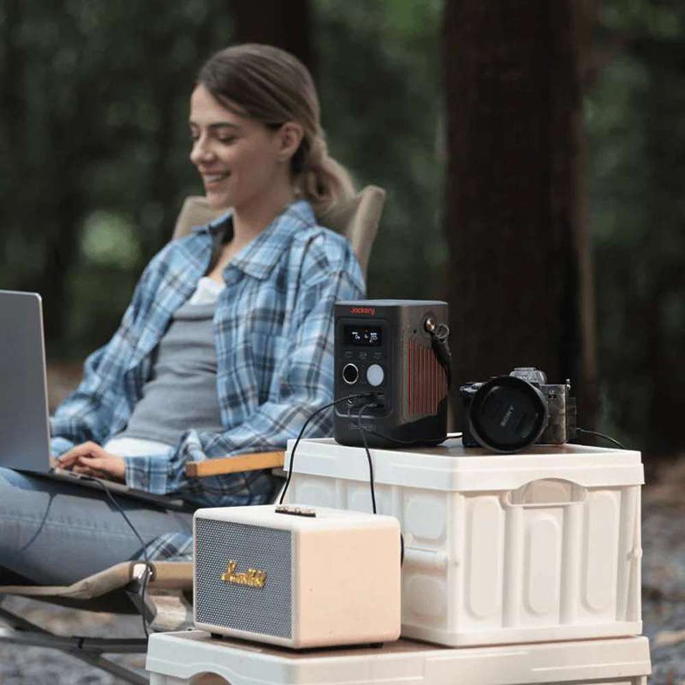 Camper uses a laptop while Jackery Explorer 300D Portable Power Station powers devices on a campsite table