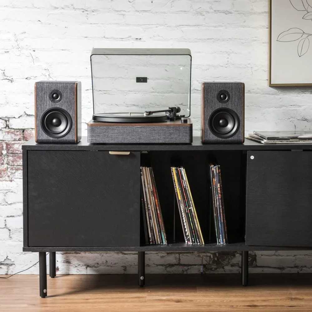 Centered on a black media cabinet with vinyl records stored below, the Crosley Nocturne Record Player With Speakers - Charcoal stands out with its matching speakers