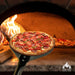 Chicago Brick Oven 7 Diameter Turning Perforated Pizza Peel holding a freshly baked pizza in front of a wood-fired oven