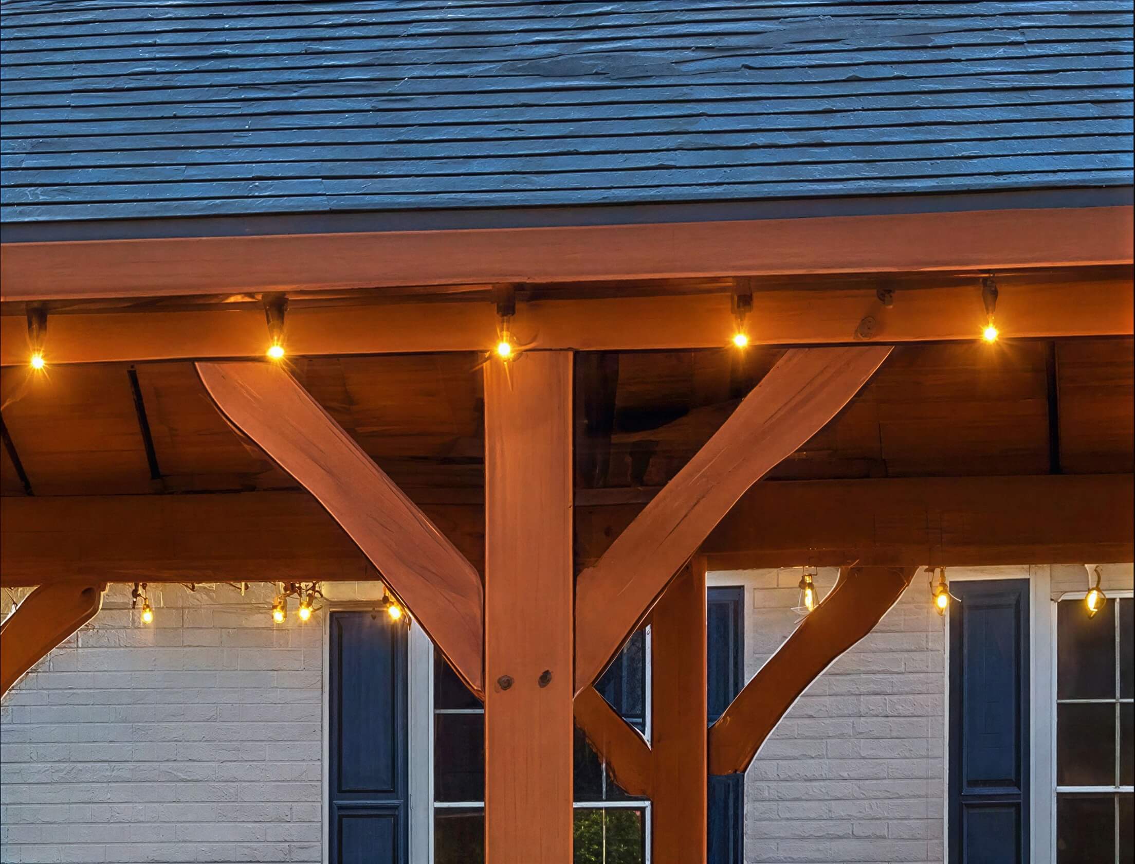 Close-up of Brookside Timber Frame Timber Crest Pavilion shows sturdy wooden beams, angled support braces, and soft string lights hanging beneath the roof
