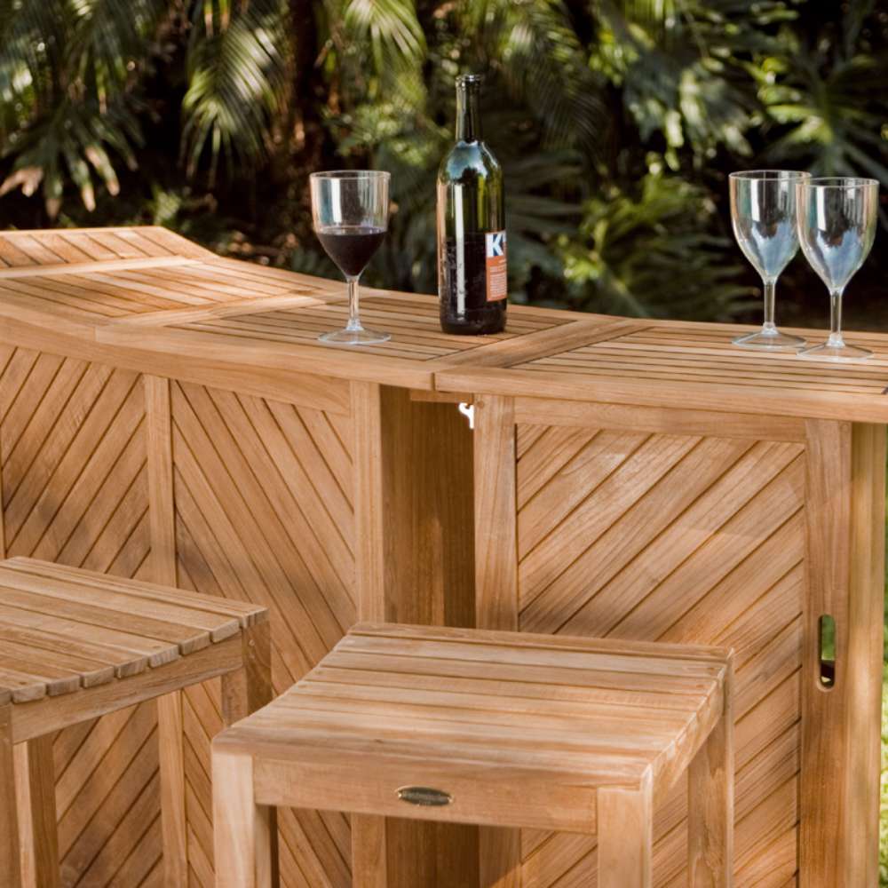 Close-up shows Westminster Teak Somerset Backless Bar Stool next to a teak bar table with a wine bottle and glasses, set against a leafy outdoor background