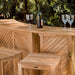 Close-up shows Westminster Teak Somerset Backless Bar Stool next to a teak bar table with a wine bottle and glasses, set against a leafy outdoor background