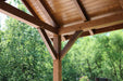 Close-up view of Brookside Timber Frame Forest Retreat Pavilion highlights its sturdy wooden beams and detailed craftsmanship against a leafy forest backdrop