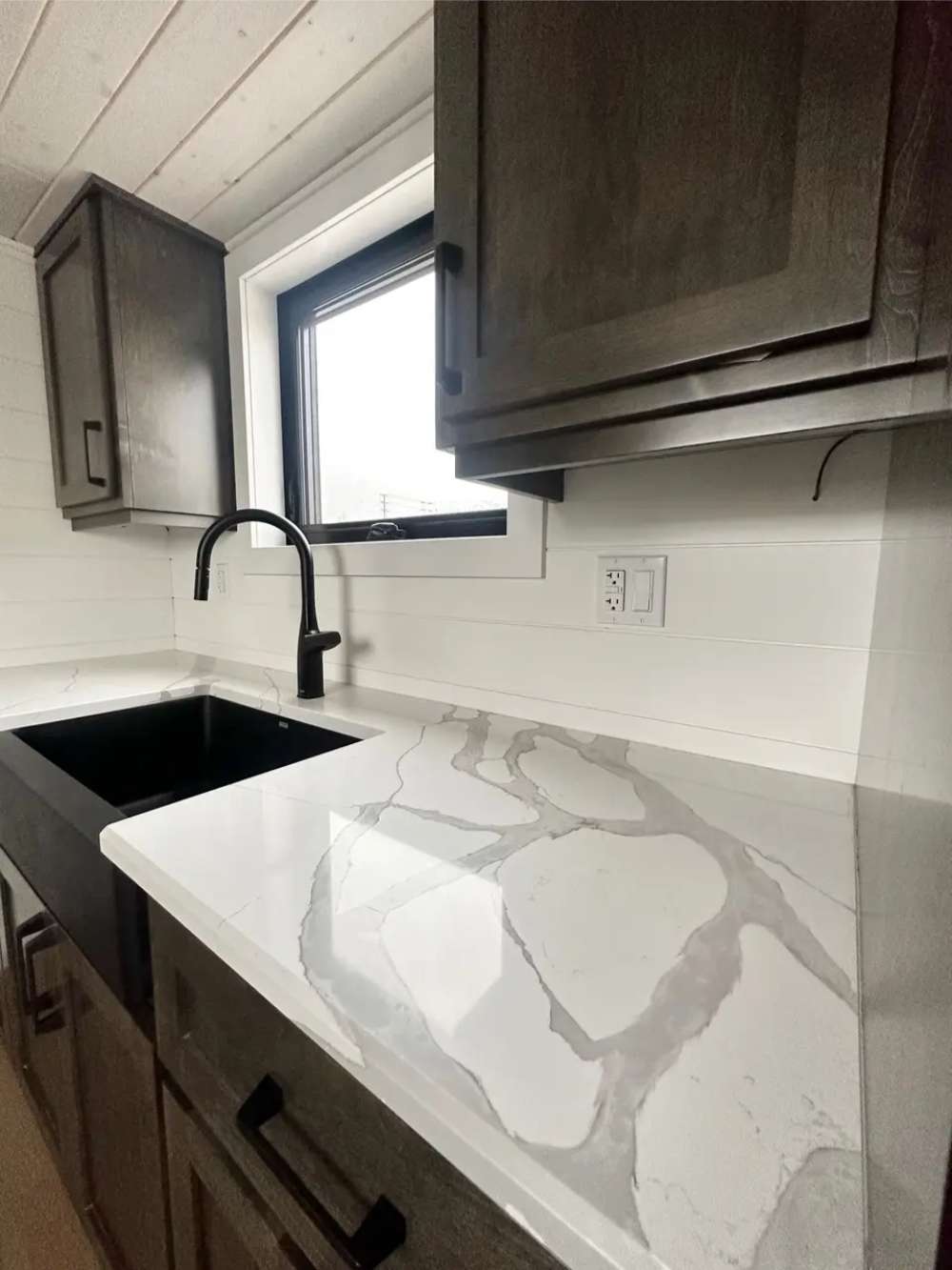 Close-up in Sawmill Structures Wanderlove Tiny Home kitchen reveals a marbled white countertop, deep black sink, and rustic dark cabinetry