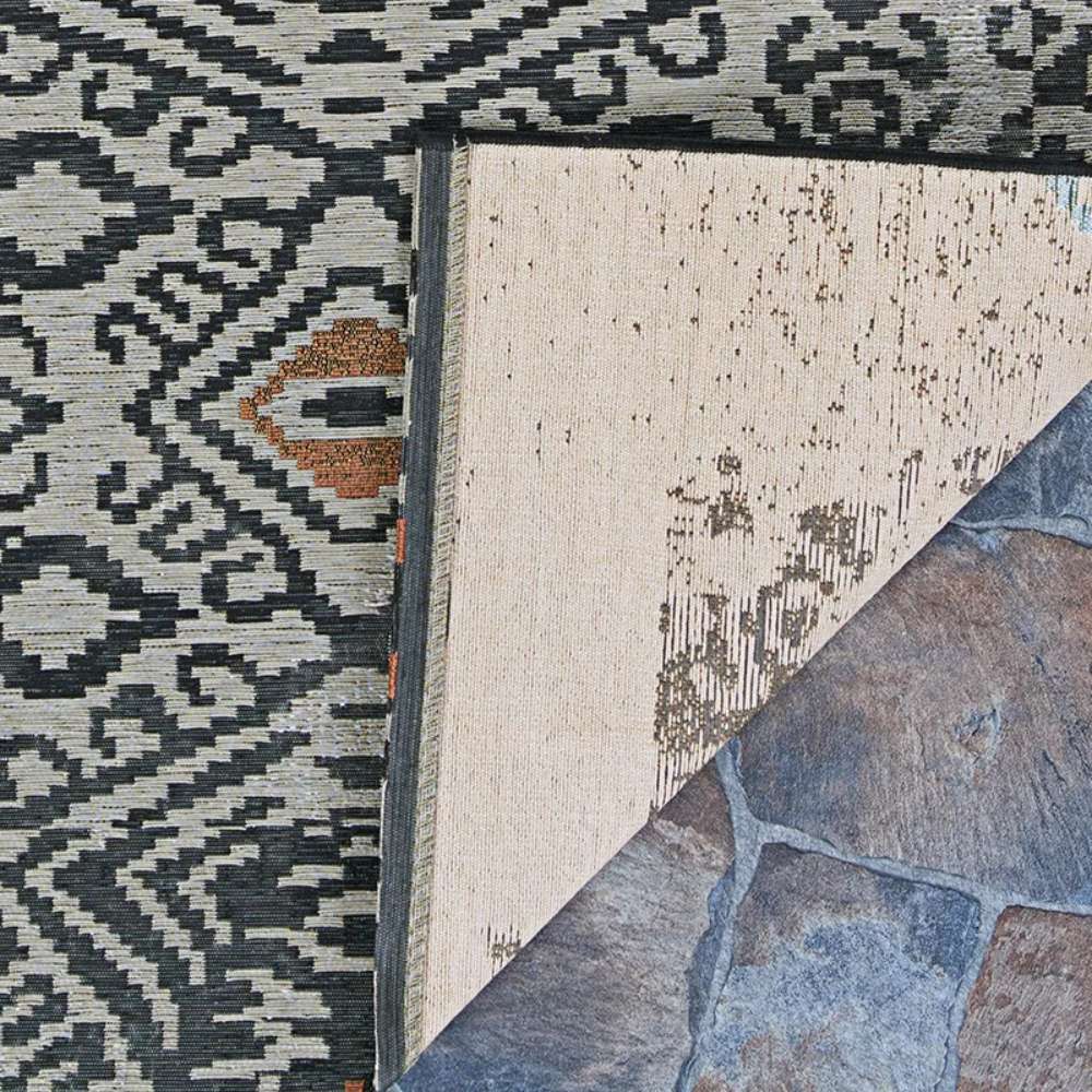 Close-up of Couristan Dolce Indoor/Outdoor Rug - Mala/Smoke shows its corner flipped over to reveal a woven beige backing against a stone floor