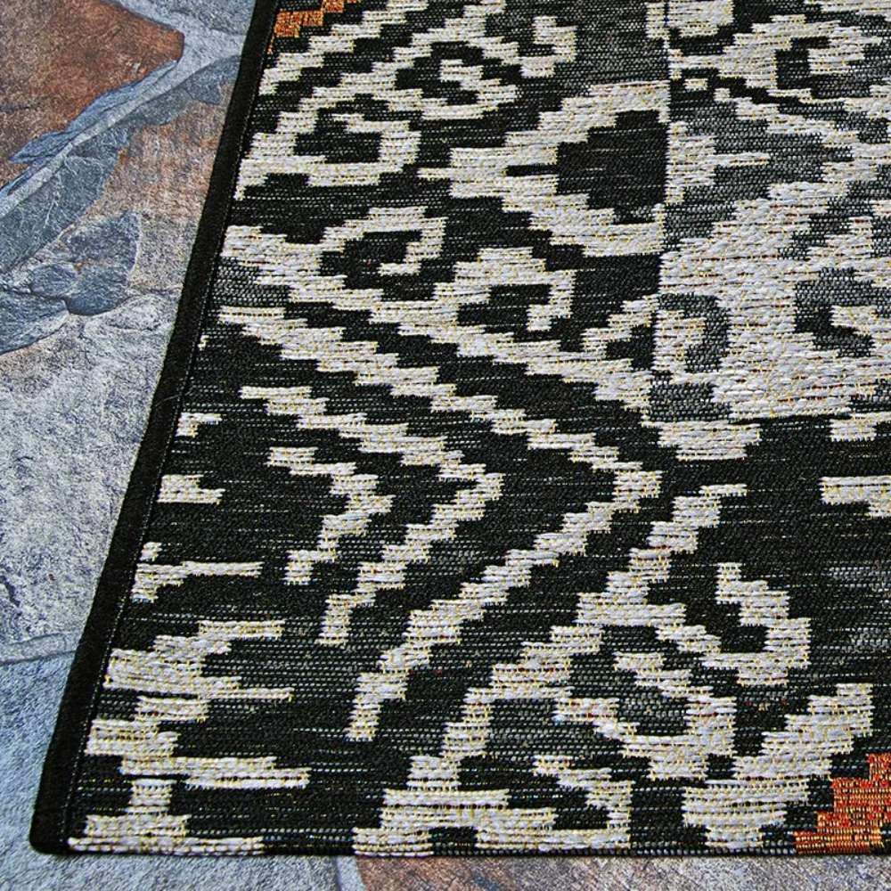 Corner of Couristan Dolce Indoor/Outdoor Rug - Mala/Smoke highlights its crisp black edge and tightly woven texture on a multicolored tile surface