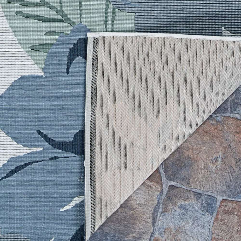 Corner of Couristan Dolce Indoor/Outdoor Rug - Novella/Grey folded back, revealing its tightly woven underside over a stone patio