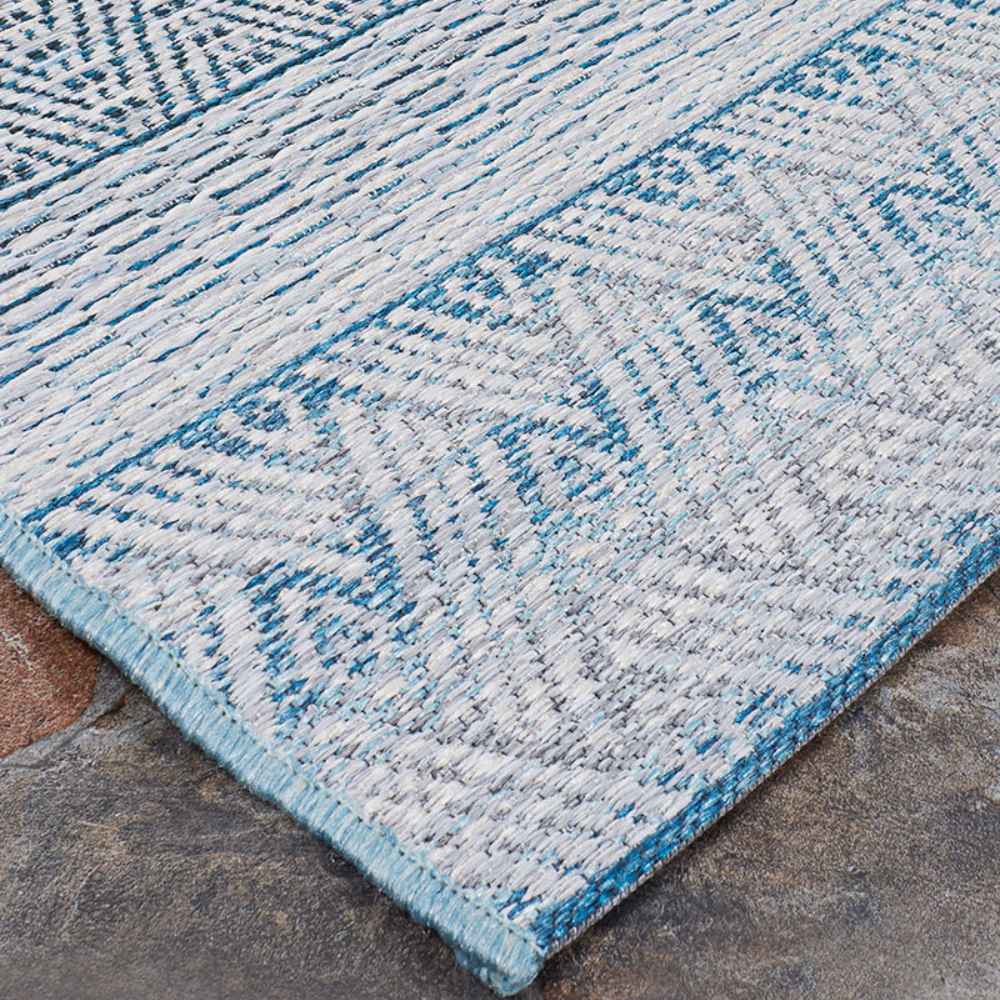 Couristan Cape Indoor/Outdoor Rug - Gables/Surf is partially rolled back on a stone surface to show off its tidy stitching and pattern consistency