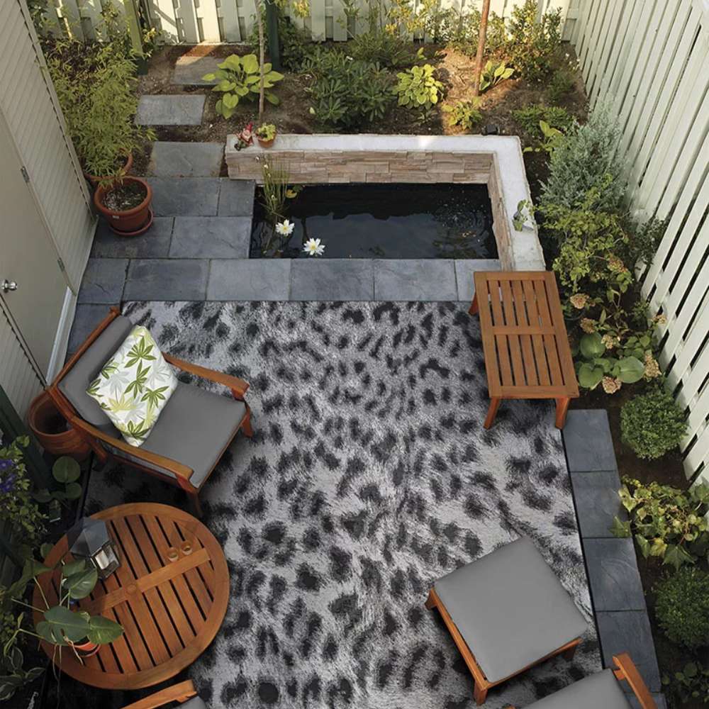 Couristan Dolce Indoor/Outdoor Rug - Lynx/Ivory-Charcoal is laid out on a cozy patio with wood furniture and greenery surrounding a small pond