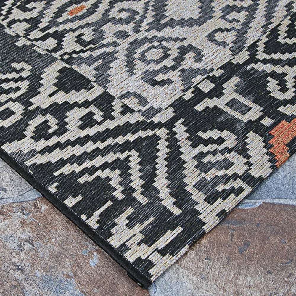 Couristan Dolce Indoor/Outdoor Rug - Mala/Smoke lies flat with sharp, tribal-inspired black and ivory patterns and touches of rust color on a stone patio