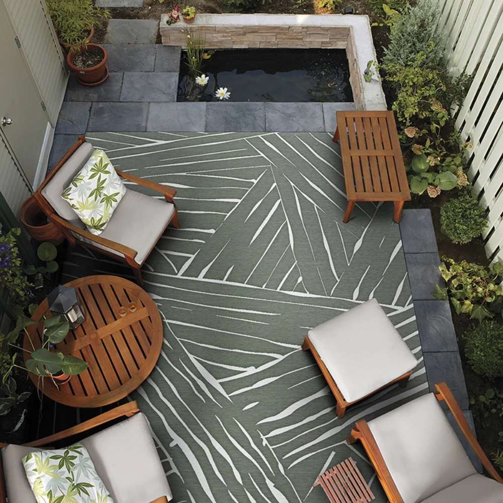Couristan Dolce Indoor/Outdoor Rug - Parquet/Fern is placed in an outdoor seating area surrounded by wood chairs and a koi pond
