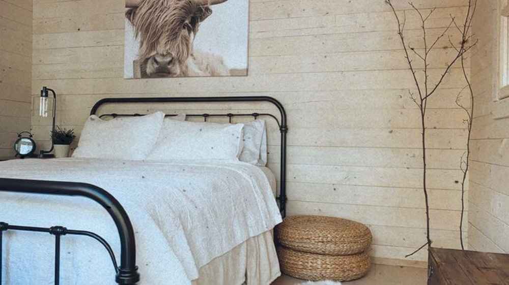 Cozy bedroom with a black metal bed frame, white bedding, and a wall-mounted animal portrait inside the Sawmill Structures Harvest Grand Bungalow 107 Garden Room