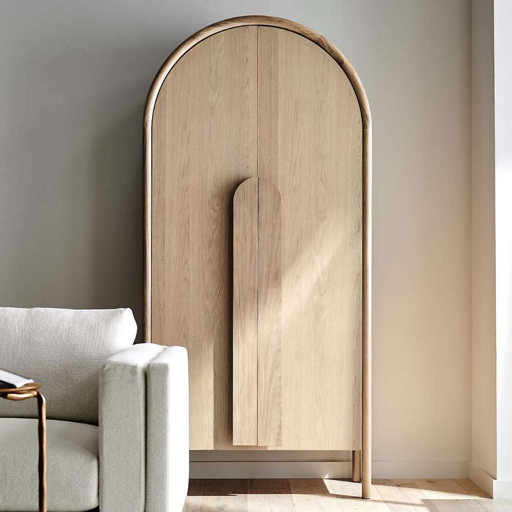 Crate & Barrel Annie 38" Natural Storage Cabinet placed beside a sofa, highlighting its tall arched design and natural wood finish