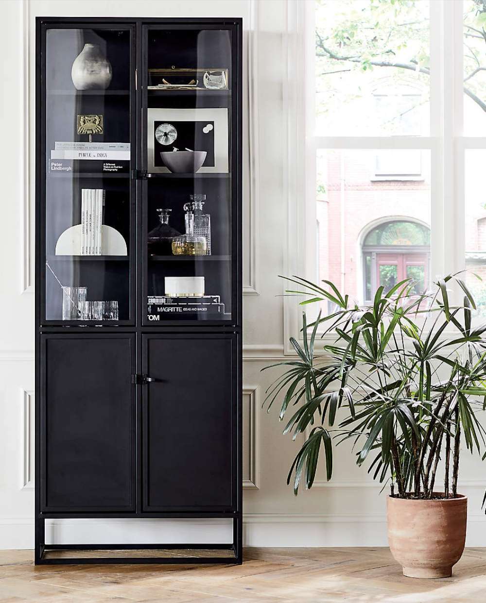 Crate & Barrel Casement 32" Black Storage Cabinet stands beside large windows, with glass shelves displaying books, glassware, and decorative objects above closed lower storage