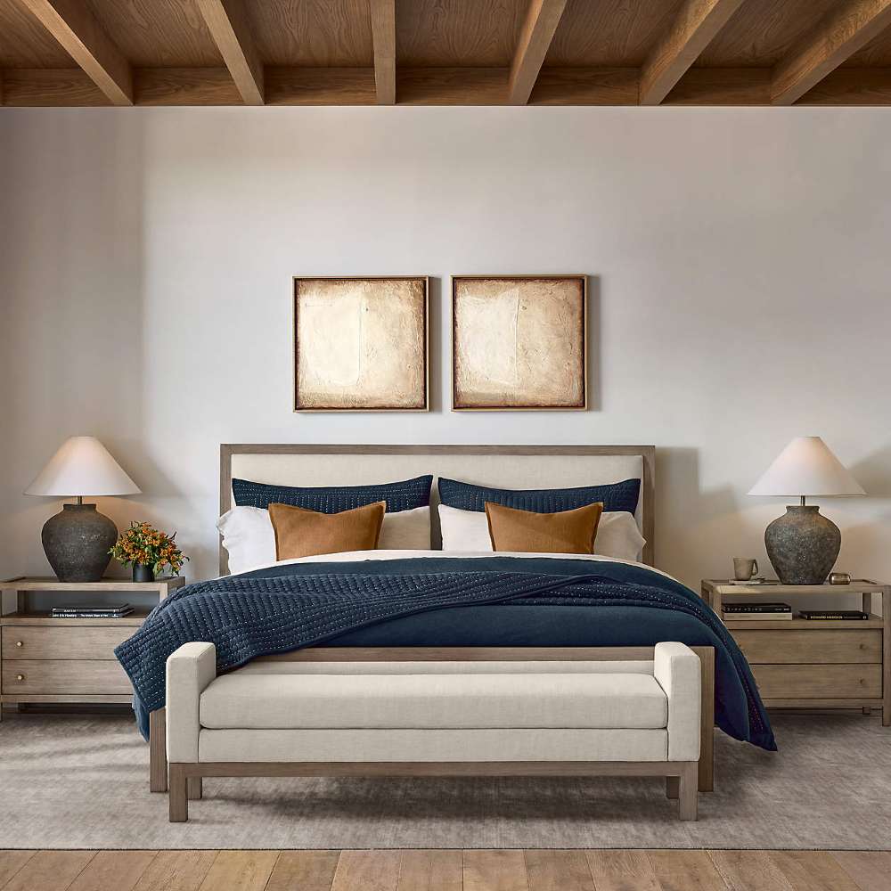 Crate & Barrel Keane Weathered Natural Wood Upholstered Queen Bed is shown in a warm bedroom with layered bedding, matching furniture, and an upholstered bench at the foot
